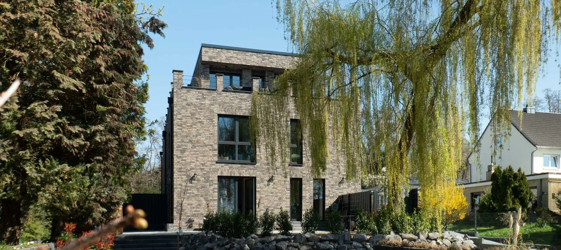 Doppelhaus BS21 mit grauem Klinker und Dachterrasse hinter einer Trauerweide